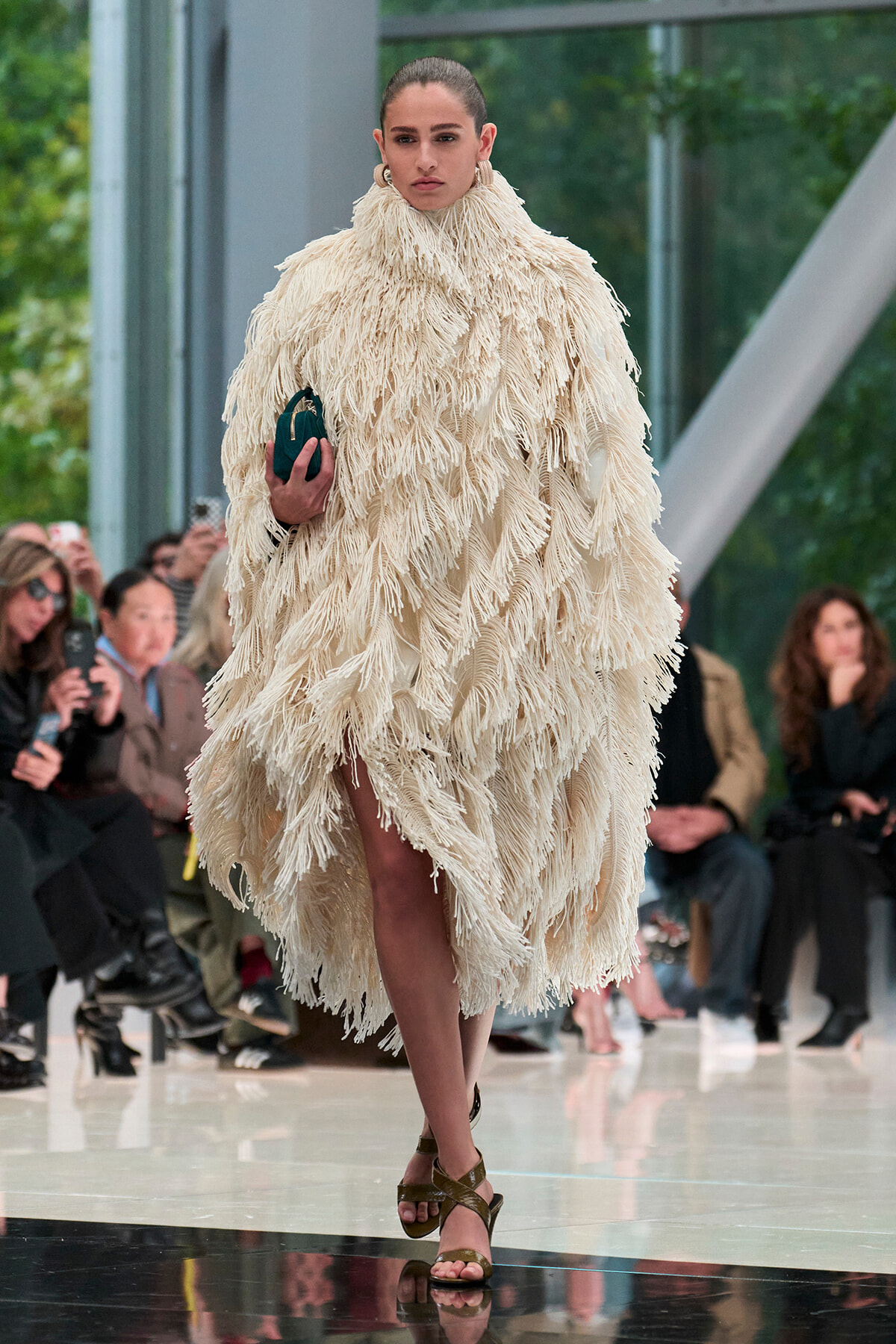 Runway model in a voluminous cream fringed coat, holding a small teal clutch, with spectators in the background.