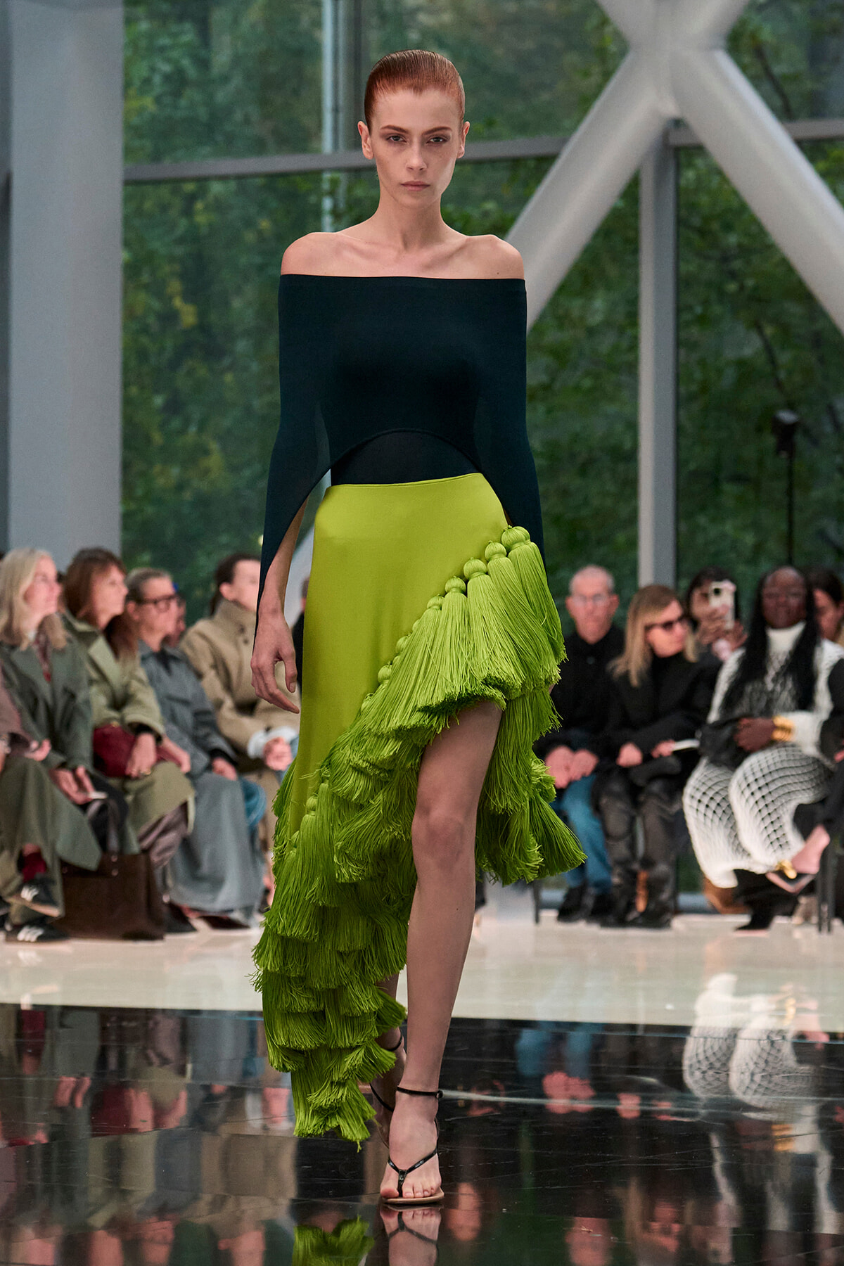 Model walks the runway in an off-the-shoulder black top paired with a lime green fringed skirt at a fashion show, audience in background