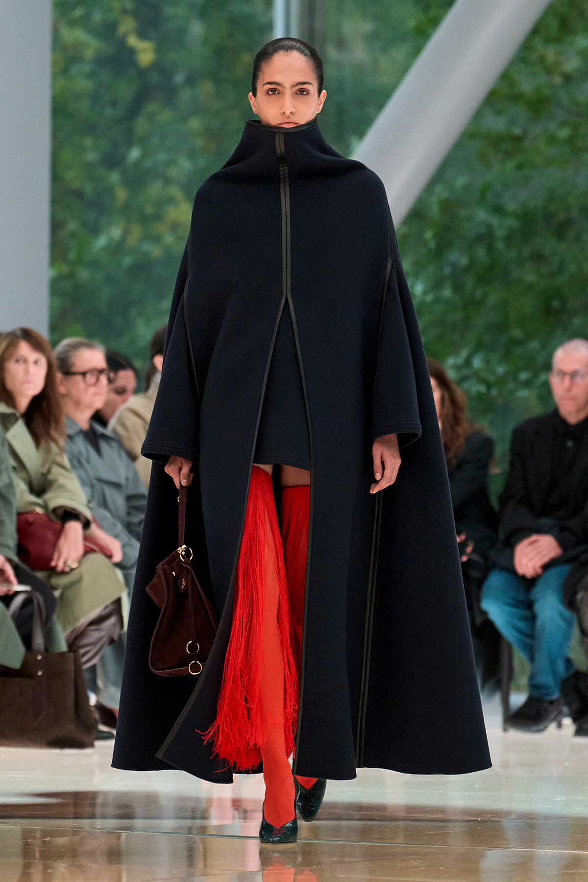 Model walks a runway in a loose navy cape with a high collar, paired with red fringe tights and a small brown handbag.