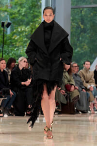Model on a runway wearing an oversized black coat with a dramatic shawl collar and fringe at the hem, plus gold sandals.