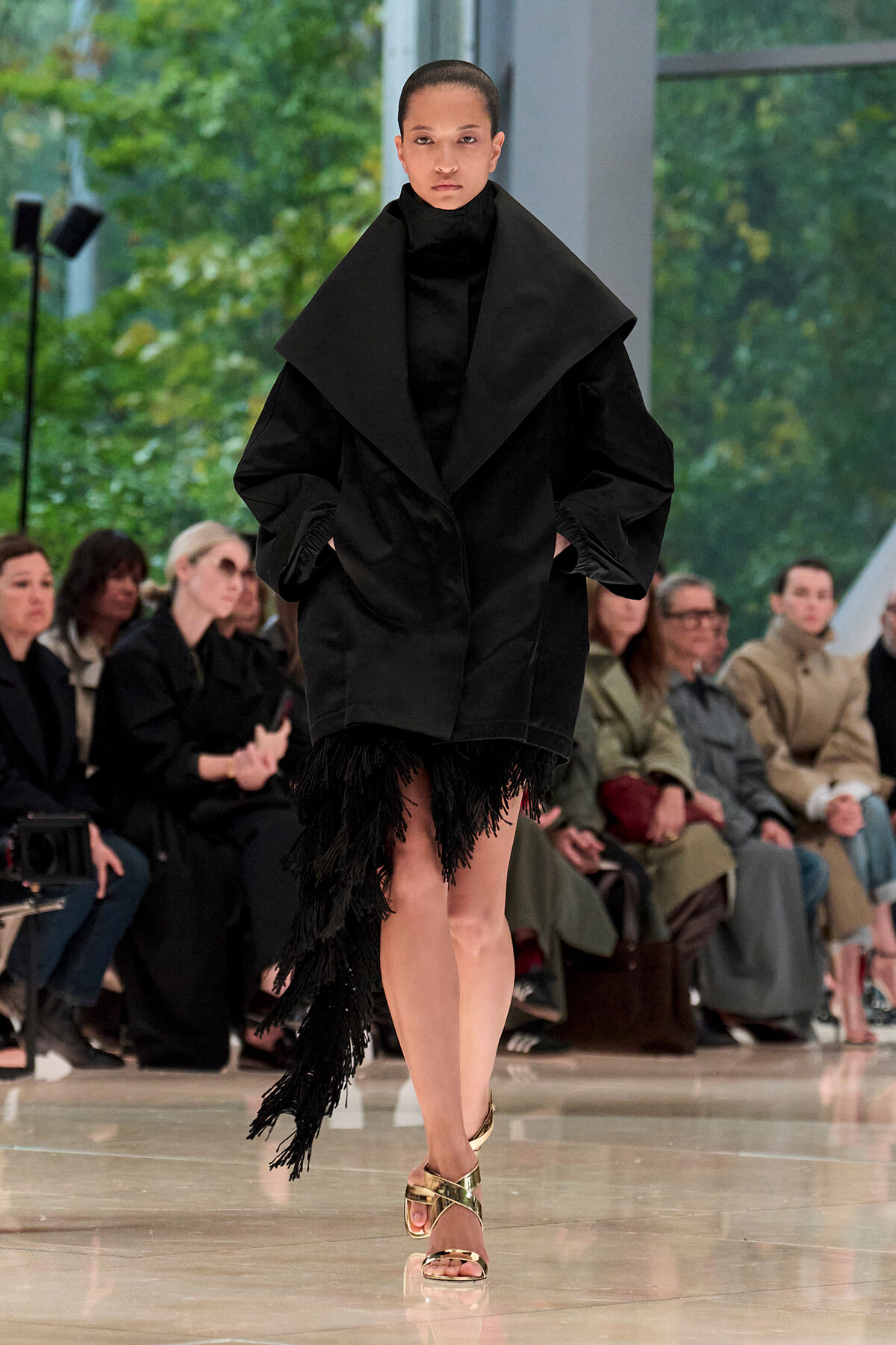 Model on a runway wearing an oversized black coat with a dramatic shawl collar and fringe at the hem, plus gold sandals.