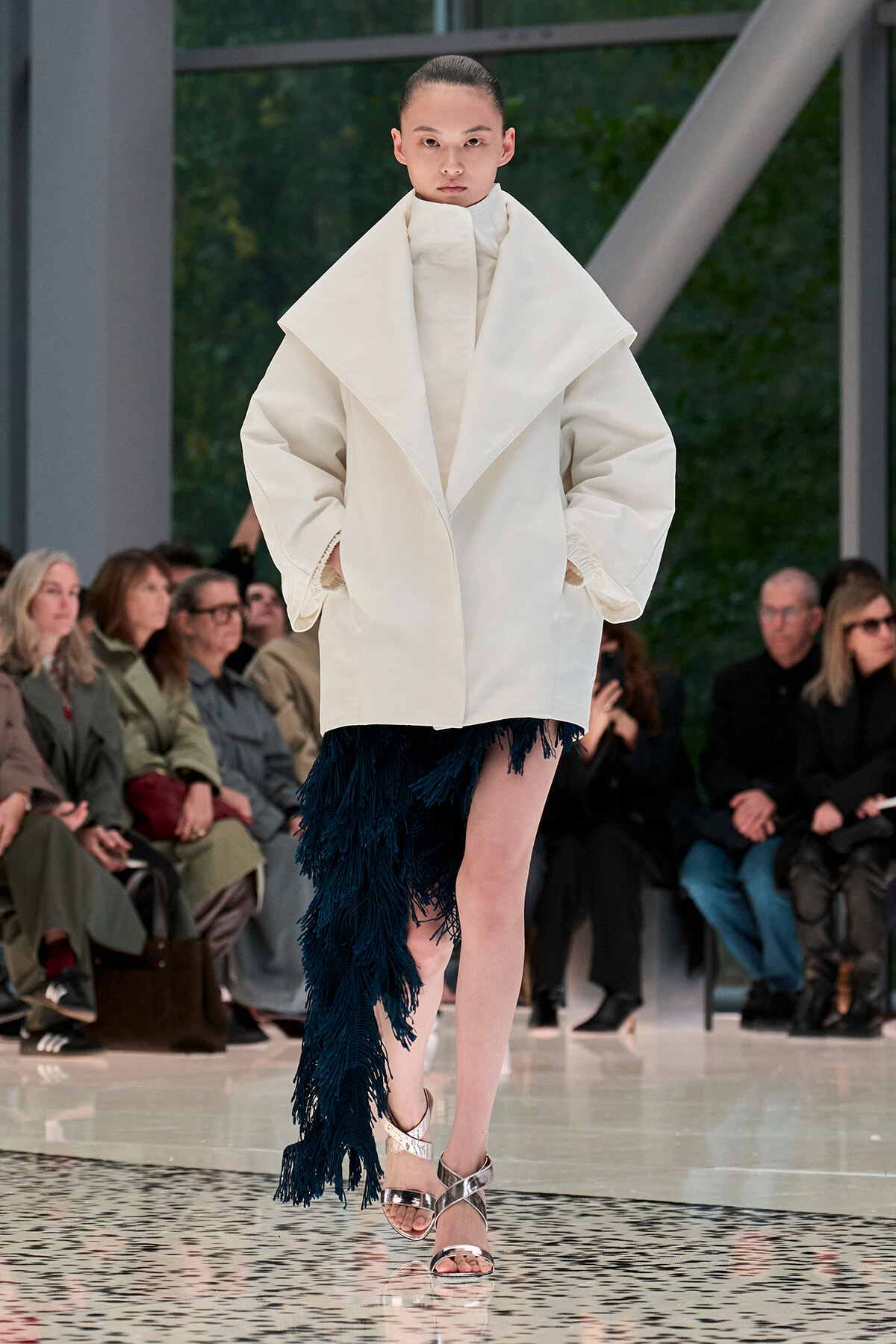 Model on a runway in a oversized white coat with a high collar and a blue fringe skirt, wearing silver sandals; audience watches in background.