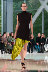 Model walks the runway in a sleeveless black high-neck dress paired with a lime-green satin asymmetric skirt; audience watches in the background.