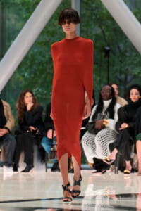 Model walks the runway in a long red form‑fitting dress with a rounded seam, black strappy heels, and a small handbag; audience seated in background.