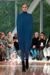 Model walks a fashion runway in a loose cobalt blue high-neck dress with gathered cuffs, blue leggings, and black ankle boots; blurred audience in background.