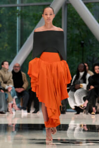Model walks the runway in a black off-the-shoulder top paired with a bright orange, layered draped skirt; spectators sit along the catwalk behind her.