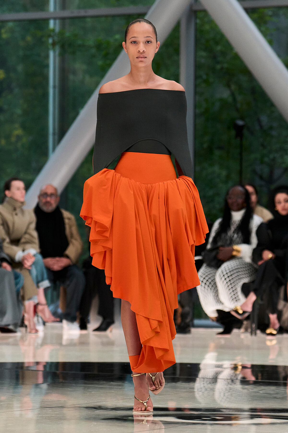 Model walks the runway in a black off-the-shoulder top paired with a bright orange, layered draped skirt; spectators sit along the catwalk behind her.