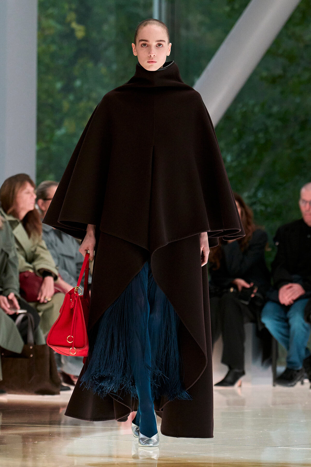 Model on a runway wearing a voluminous black cape with a high neck, over a blue fringed skirt, teal tights, and metallic silver shoes, carrying a red handbag.