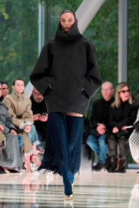 Model walks the runway in an oversized black coat with a high collar, paired with a blue fringed skirt and gold shoes.