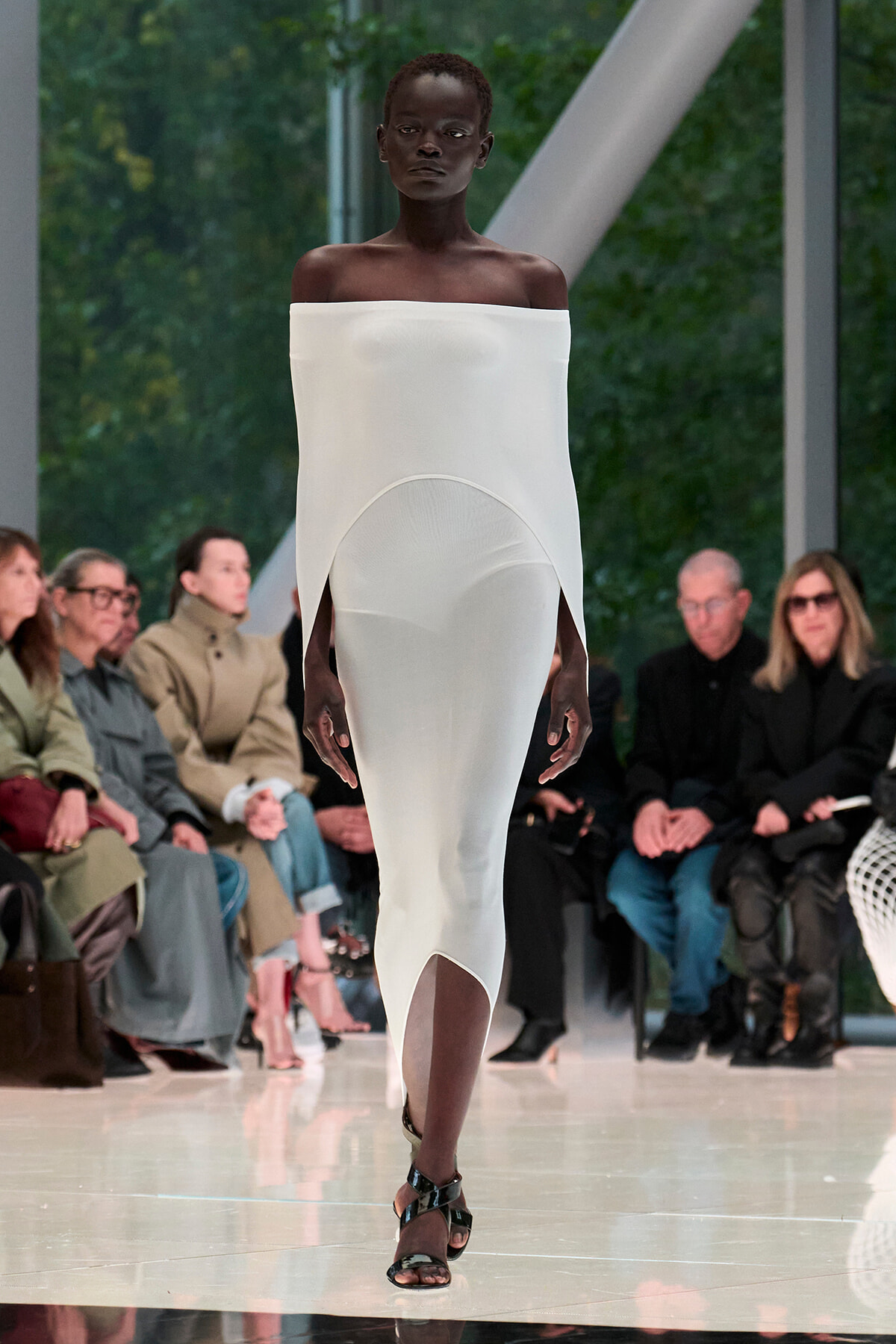 Black model on a runway wearing a white off-the-shoulder column dress with a front slit, audience visible in the background.