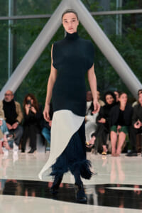 Model walks a runway in a black sleeveless turtleneck dress with a white asymmetric panel and navy fringe at the hem, audience watching in the background?