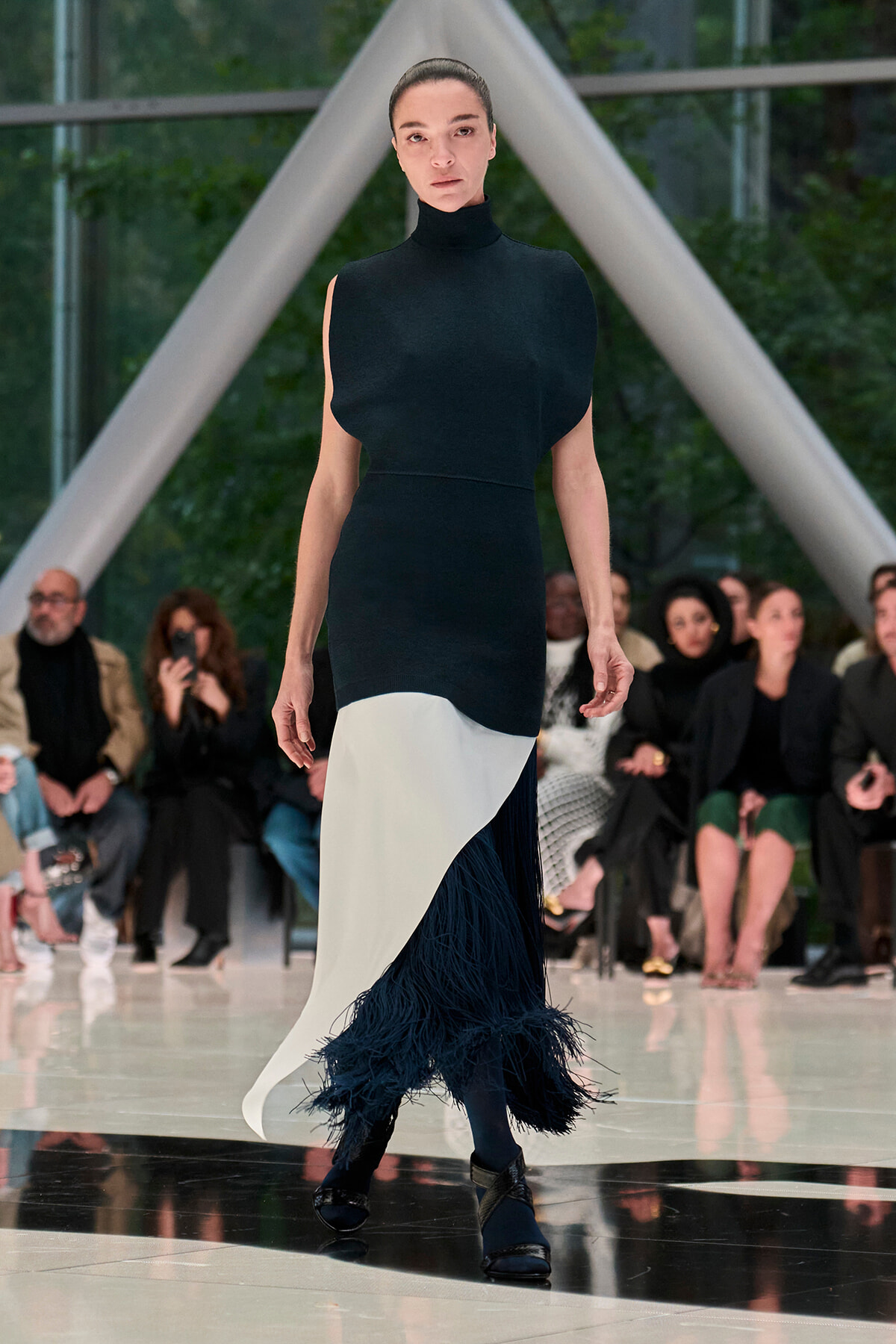 Model walks a runway in a black sleeveless turtleneck dress with a white asymmetric panel and navy fringe at the hem, audience watching in the background?