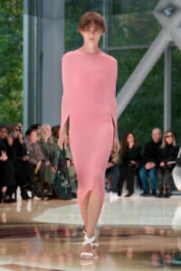 Model in a pastel pink midi dress with sculptural curved panels, carrying a dark green handbag, and wearing white sandals on a runway.