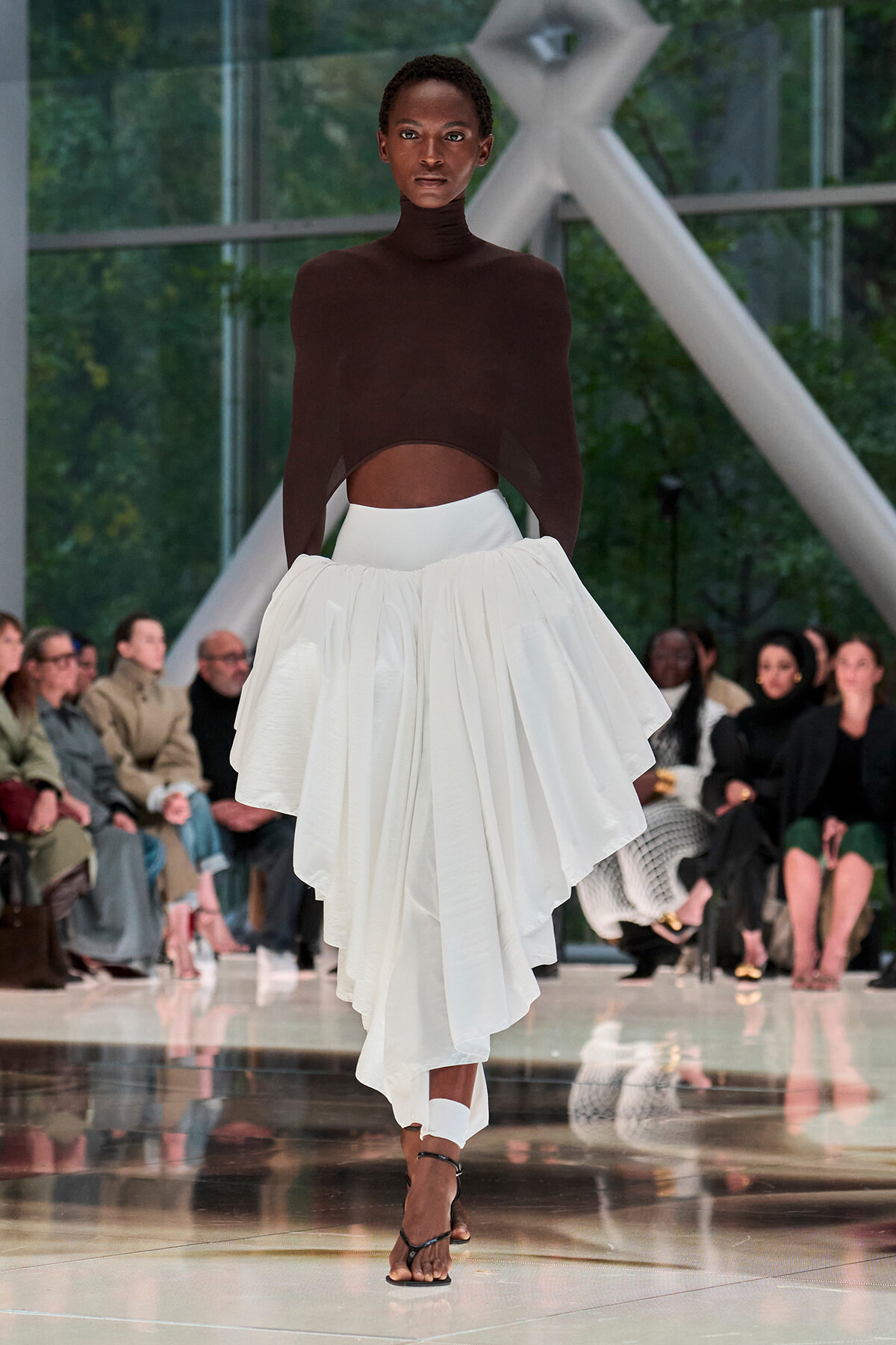 Model walking a fashion runway in a dark brown turtleneck crop top and a voluminous white asymmetric skirt, with spectators in the background.