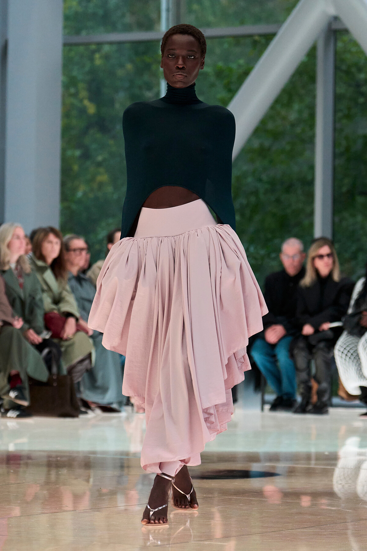 Model walking a fashion runway in a black turtleneck top and voluminous pale pink skirt, with audience watching in the background