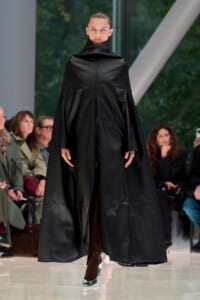 Model walks the runway in a long black leather cape with a high collar, audience watching.