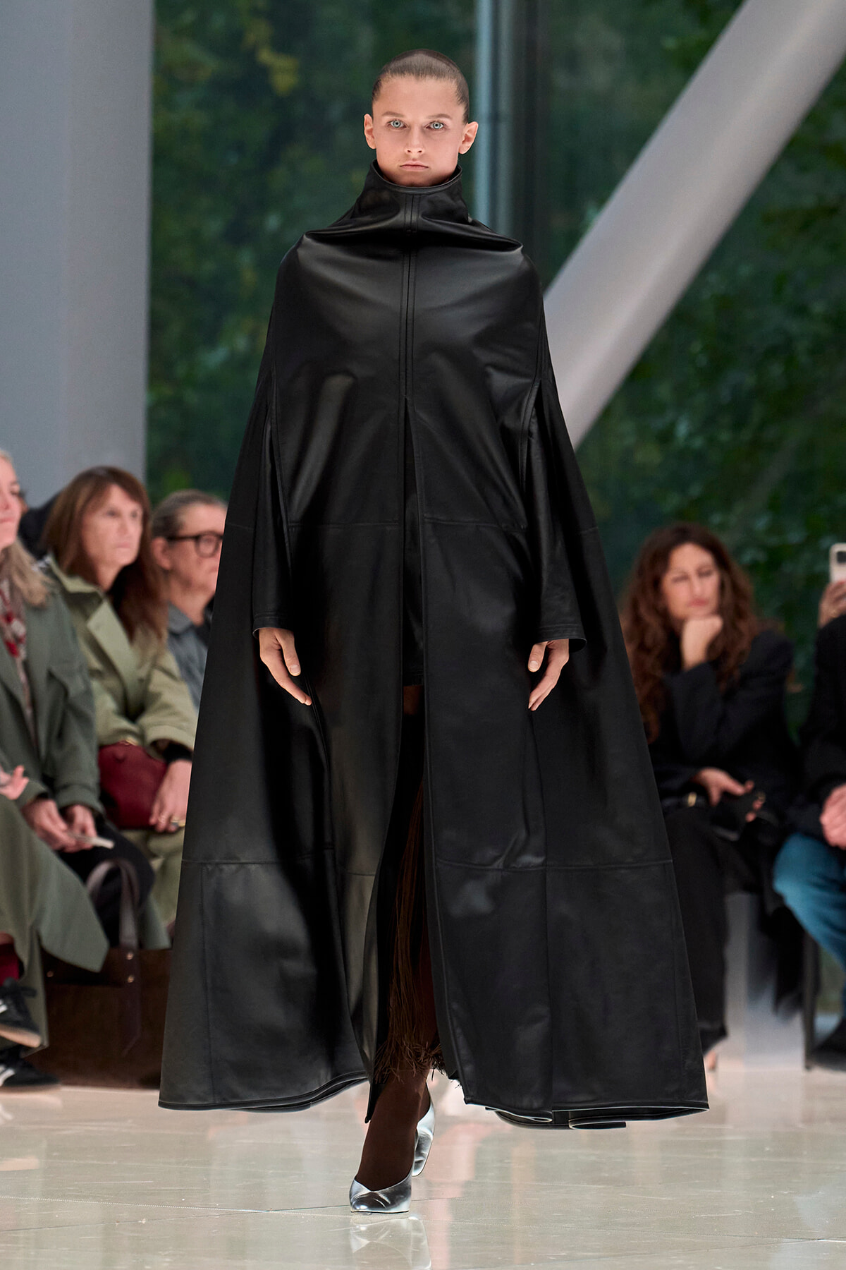 Model walks the runway in a long black leather cape with a high collar, audience watching.