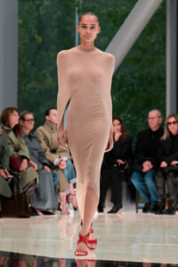 Model walking a fashion runway in a nude sheer bodycon dress with red strappy sandals; audience seated along the sides.