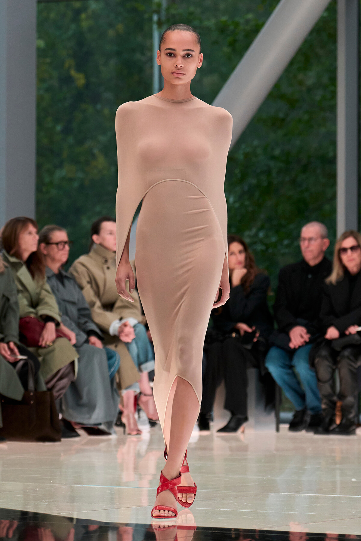 Model walking a fashion runway in a nude sheer bodycon dress with red strappy sandals; audience seated along the sides.