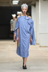 Model walks a fashion show wearing an oversized blue striped button-down shirt dress with off-shoulder sleeves and a sculptural white spiked headpiece.