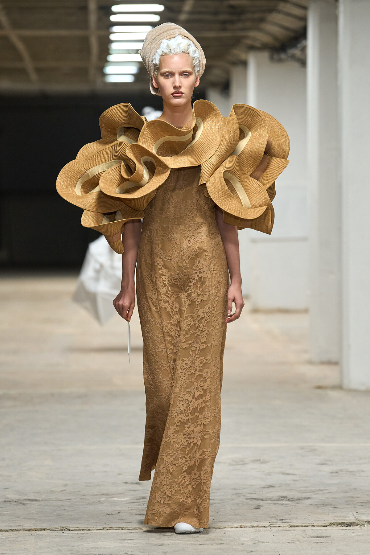 Model on a runway wearing a long beige lace gown with oversized sculptural ruffle shoulders and a wrapped headpiece.