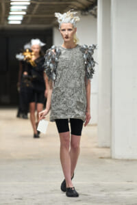 Model wearing a silver crushed-foil tunic with metallic spiky shoulder embellishments on a runway, pale blonde hair styled sculpturally in a white updo.