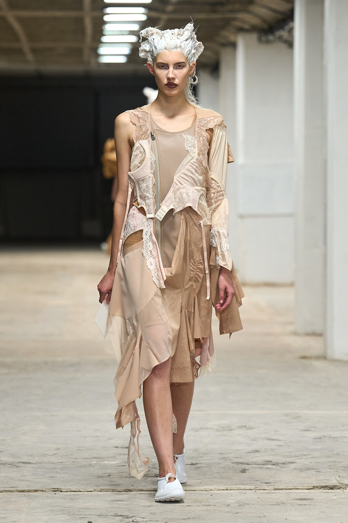 Model walking a runway in a beige patchwork dress with lace appliqués, white sneakers, and a sculpted white hairdo in an industrial venue.