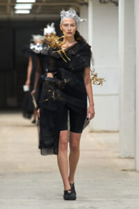 Runway model in black avant-garde dress wrapped with sheer layers, wearing a silver sculpted headpiece and gold aerodynamic shoulder accents, walking down a studio hallway.