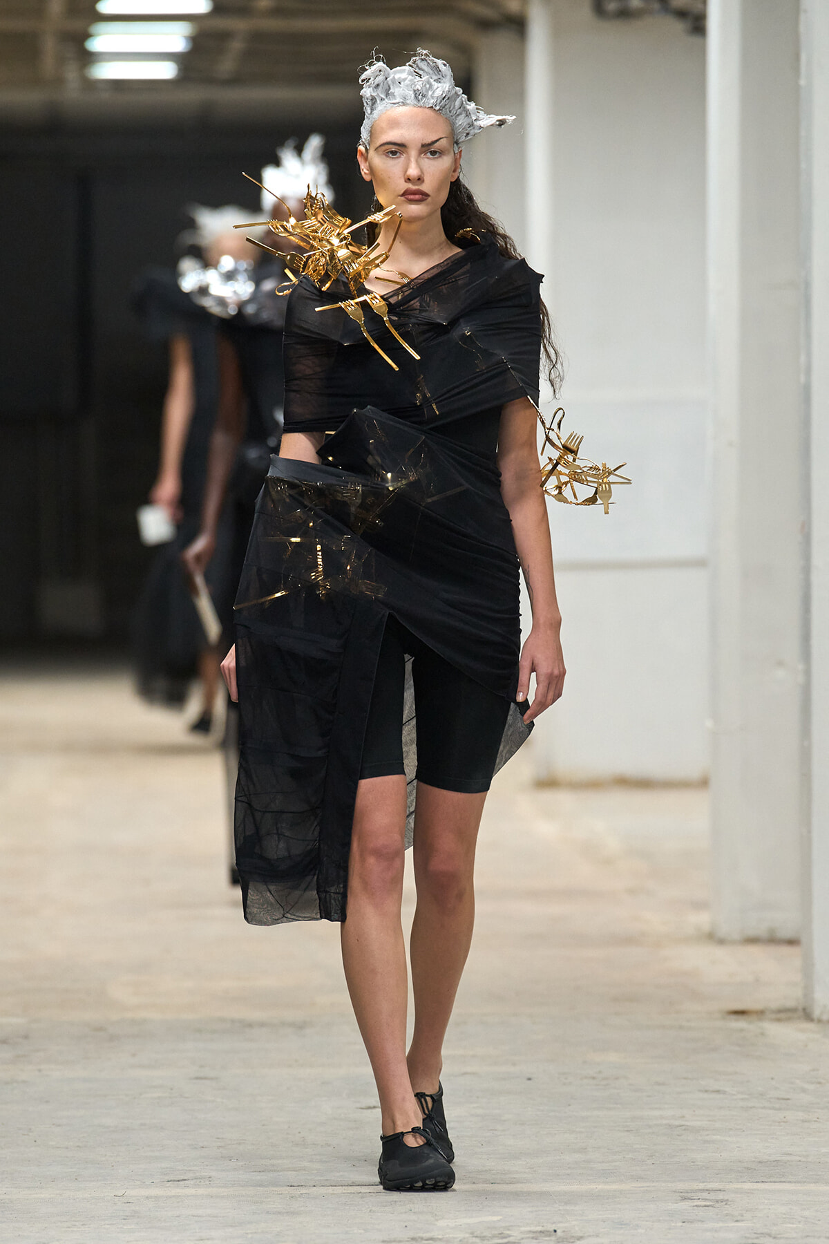 Runway model in black avant-garde dress wrapped with sheer layers, wearing a silver sculpted headpiece and gold aerodynamic shoulder accents, walking down a studio hallway.