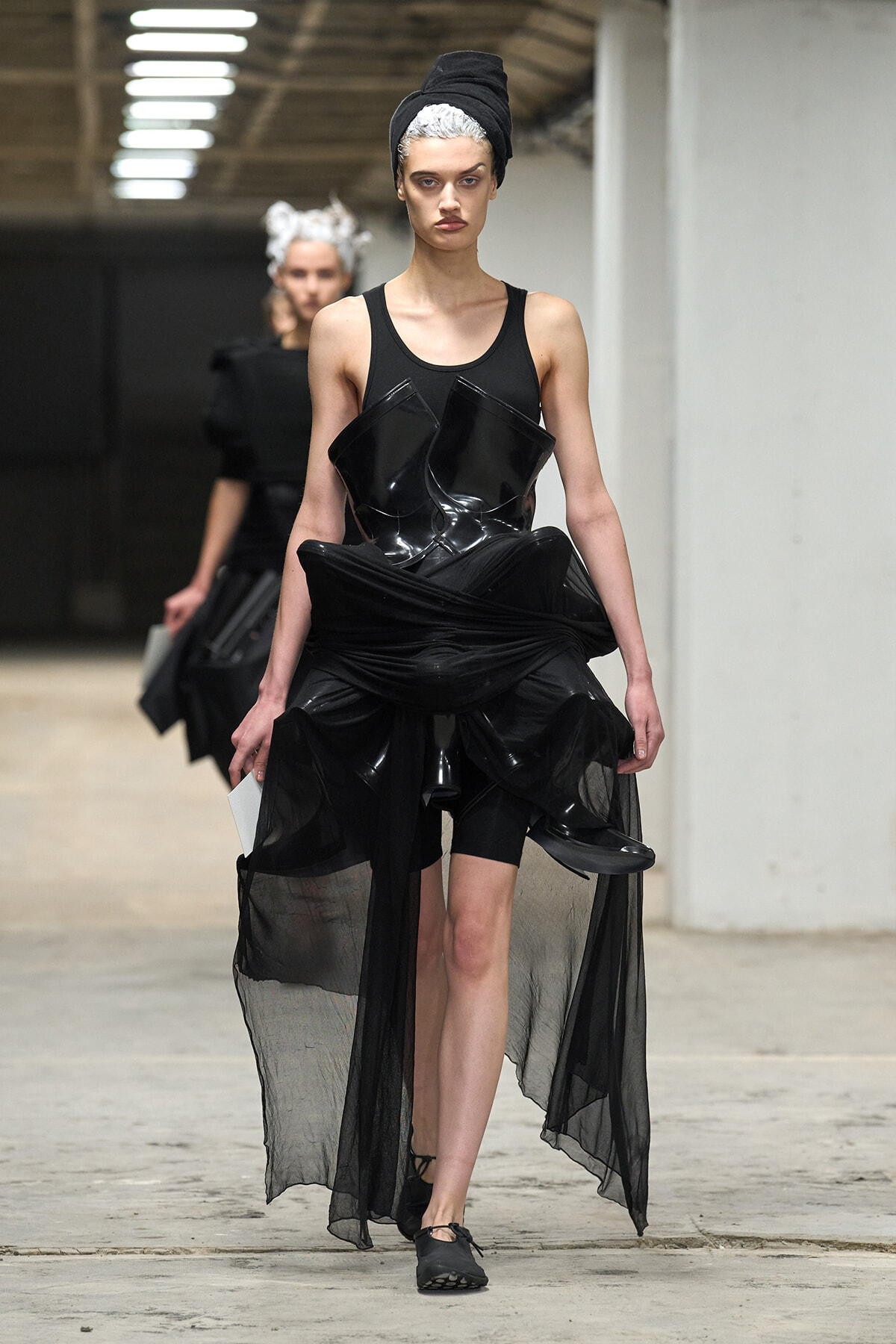 Runway look: a model in a black avant-garde outfit with a sculptural vinyl bodice and sheer draped skirt, head wrapped in black fabric.