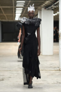 Model in a black, layered gown with a dramatic plastic shoulder collar and white sculpted hair walks a runway in an industrial space.