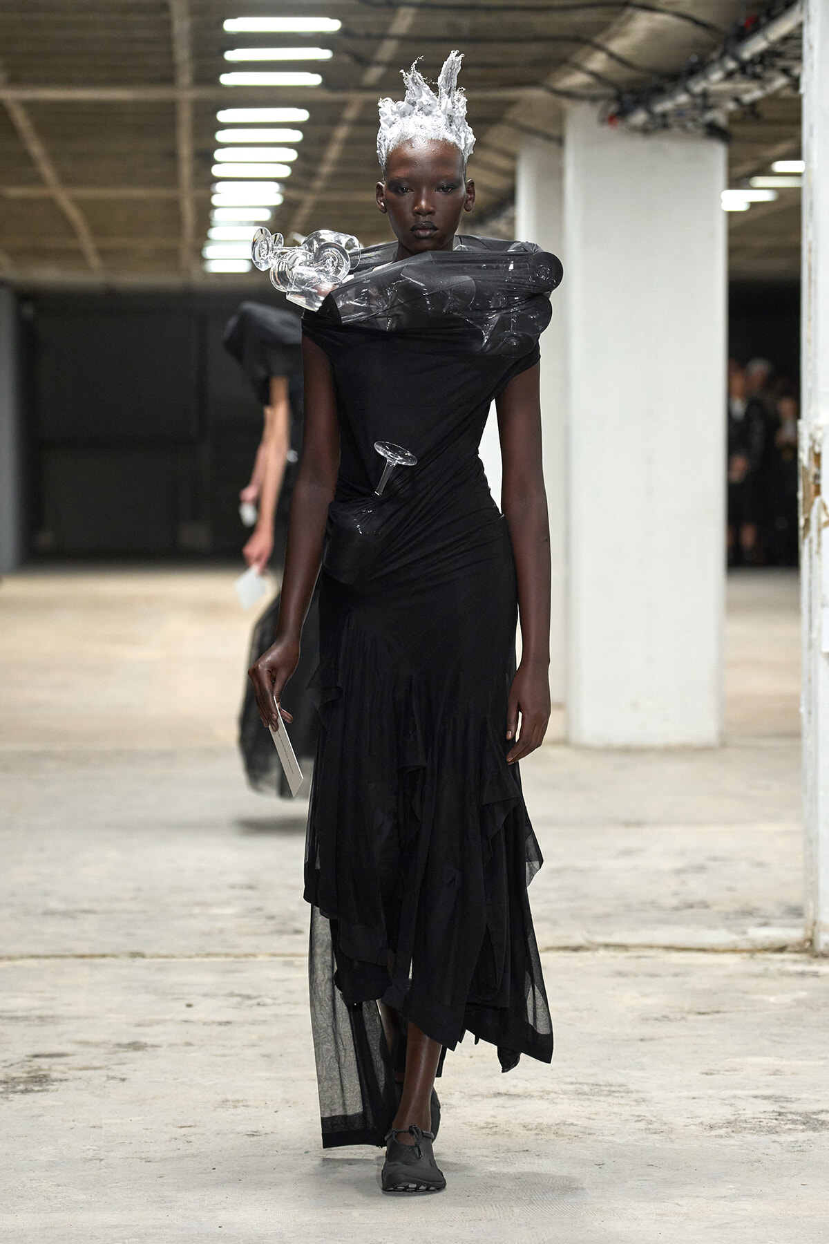 Model in a black, layered gown with a dramatic plastic shoulder collar and white sculpted hair walks a runway in an industrial space.