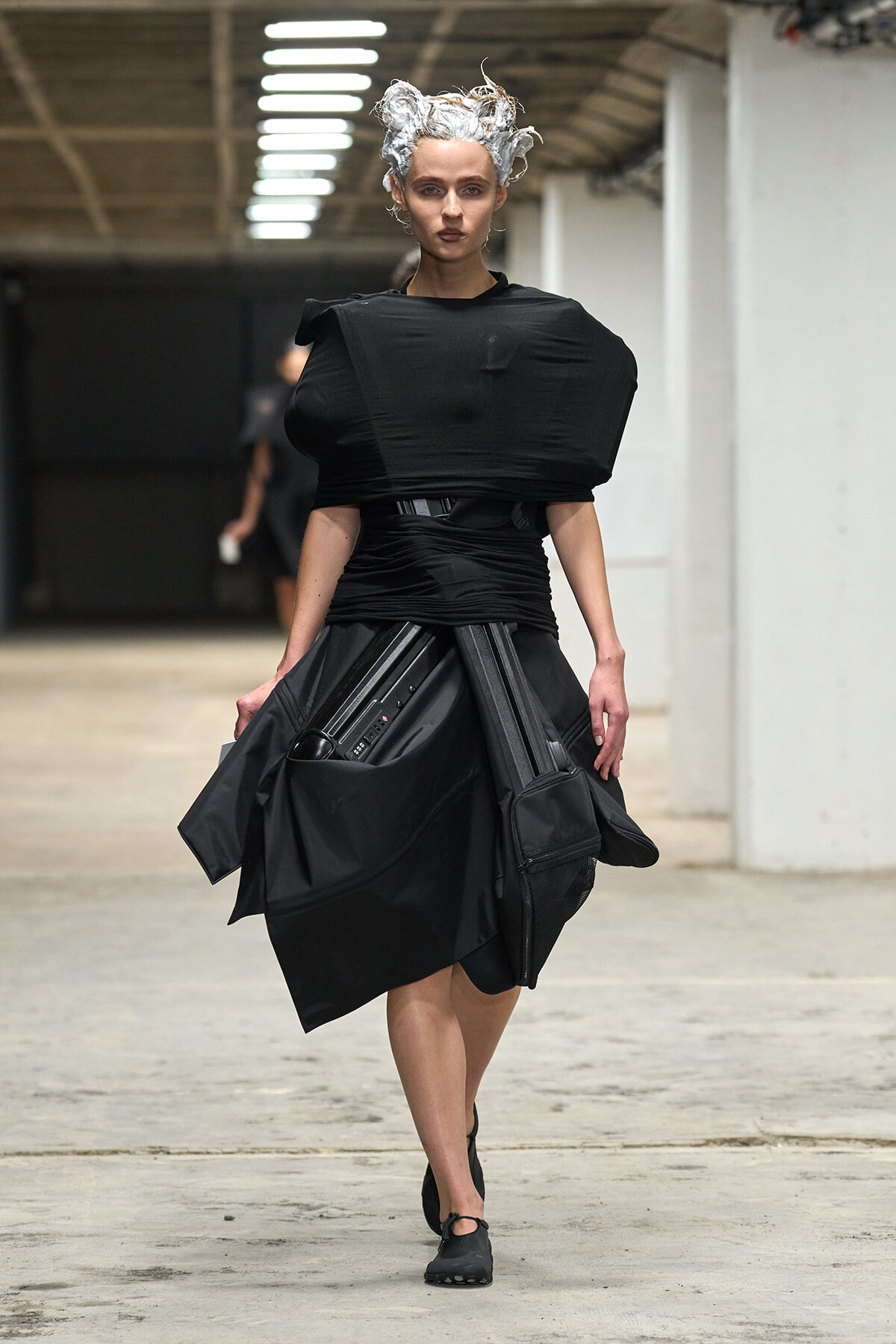 Model walking a runway in a black sculptural outfit with a voluminous top and structured, draped skirt.