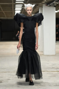 Model wearing a black, sculptural gown with oversized shoulder wings and a sheer tulle mermaid skirt on a runway.