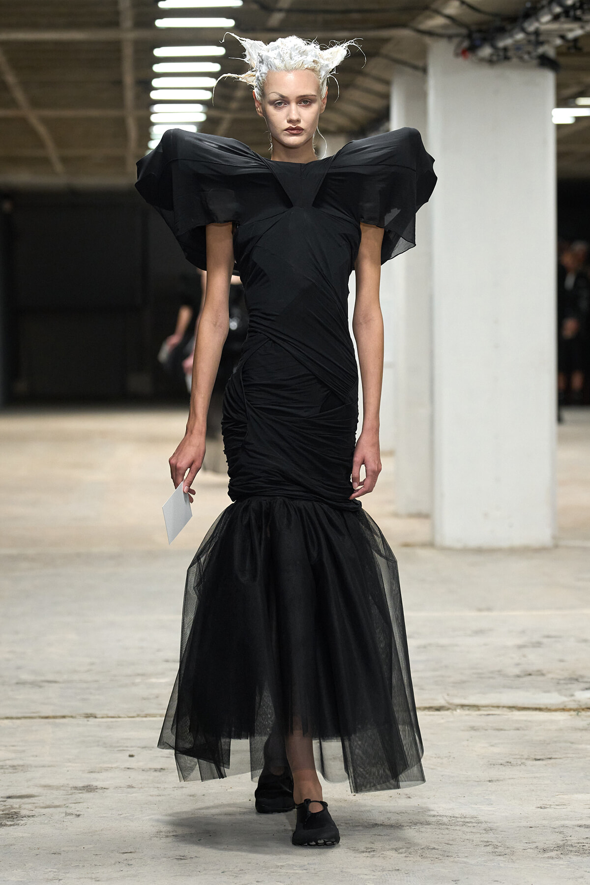 Model wearing a black, sculptural gown with oversized shoulder wings and a sheer tulle mermaid skirt on a runway.