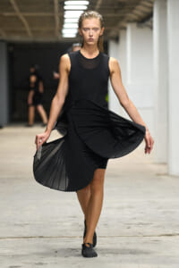 Model walking the runway in a sleeveless black dress with a flowing pleated skirt, in a backstage fashion show setting