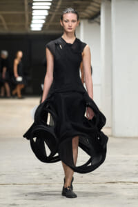 Model walking in a dramatic black dress with large circular fabric loops cascading from the skirt on a runway.