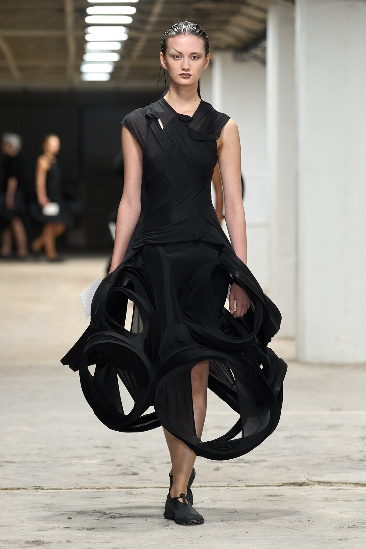 Model walking in a dramatic black dress with large circular fabric loops cascading from the skirt on a runway.