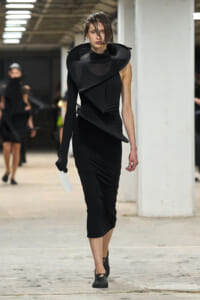 Runway model in a black sculptural dress with oversized collar and layered panels, walking in an industrial venue.