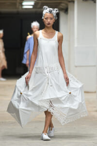 Model walks the runway in a white sleeveless dress with lace trim and an oversized, umbrella-like skirt; white shoes and a foil headpiece complete the look.