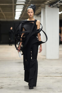 Female model on a runway wearing an avant‑garde black outfit with looping strap sculptures and a wrapped headpiece