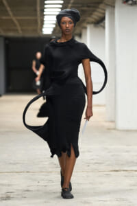 Model walking a fashion runway in a form-fitting black dress with circular fabric loops and a black headwrap.