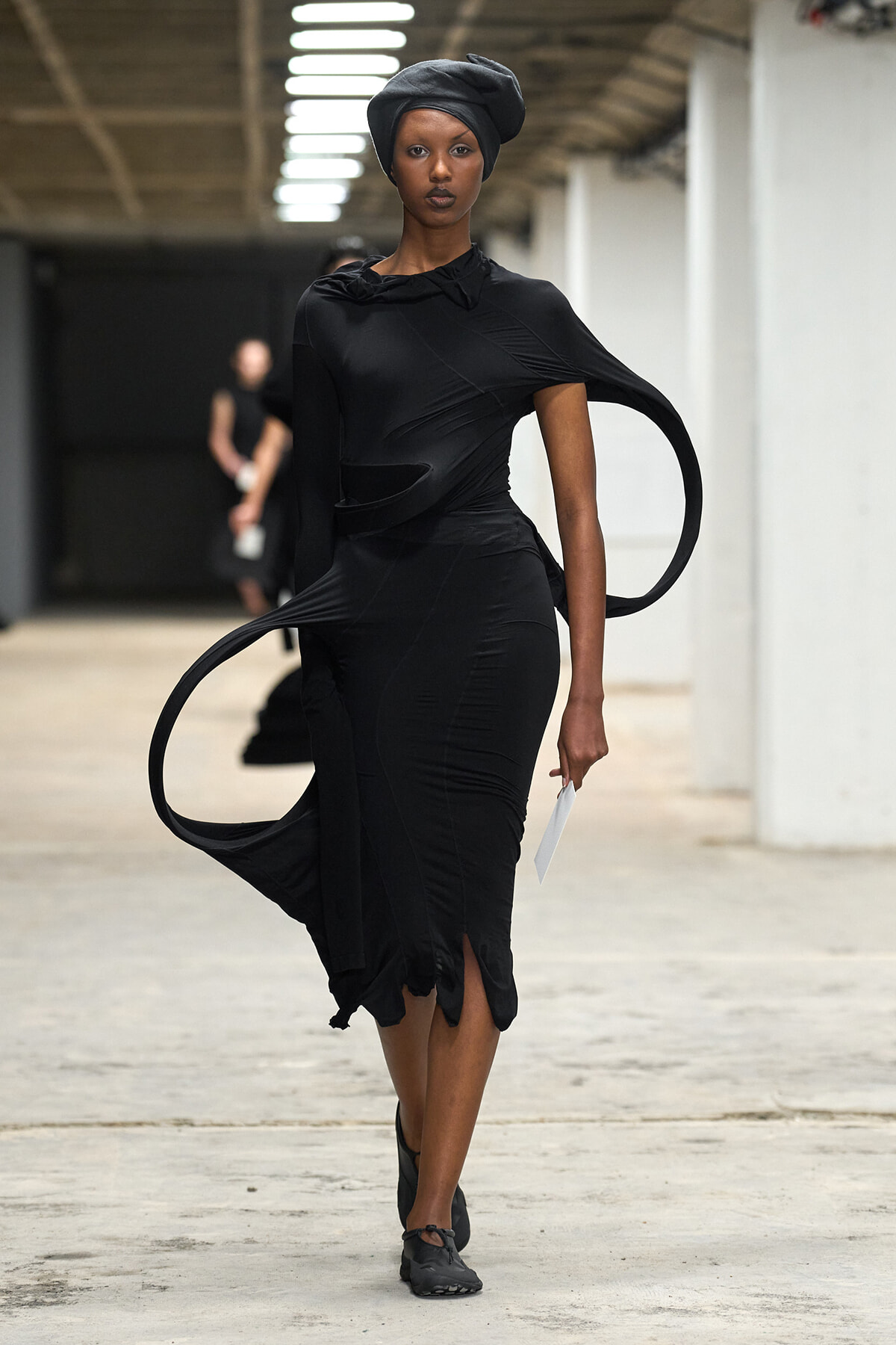 Model walking a fashion runway in a form-fitting black dress with circular fabric loops and a black headwrap.
