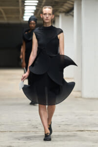 Model walking in a black avant-garde dress with large circular layered panels on a runway, holding a small white card in her left hand