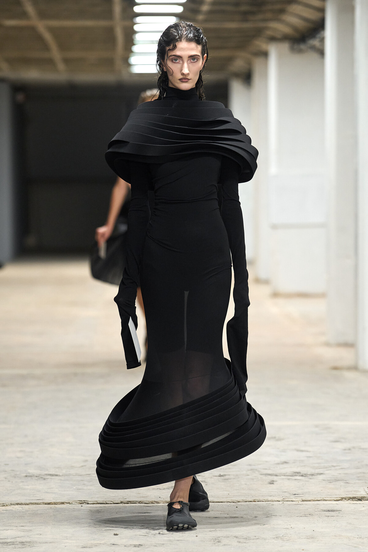 Runway model in a dramatic black sculptural gown with a multi-layered, oversized collar and swirling hem.