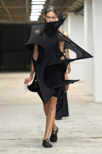 Model wearing a black, sculptural dress with cutouts and angular panels, holding a white tag as she walks in an industrial venue.