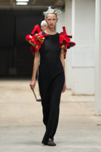 Model walking on runway in black jumpsuit with oversized red sculptural shoulder pieces reminiscent of high-fashion armor, in an industrial setting.