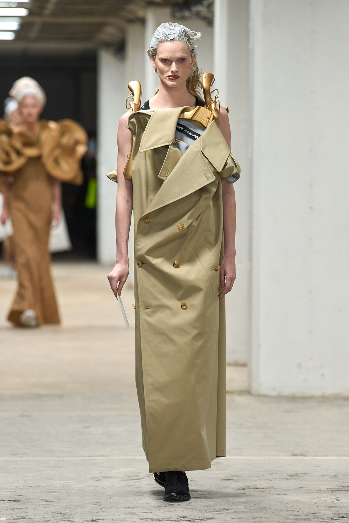 Model on a runway wearing a long beige trench dress with oversized angular lapels, gold buttons, and sculptural shoulder accents; hair wrapped in a net.