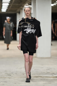 Model walking runway in an oversized black T-shirt with bold white graphic, paired with black bike shorts; a spiked chain adorns her shoulder and chest, and she wears black shoes in an industrial venue.