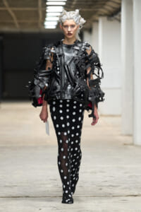 Model walking in a runway outfit: black lattice leather top over polka-dot tights, white hair, industrial backdrop.